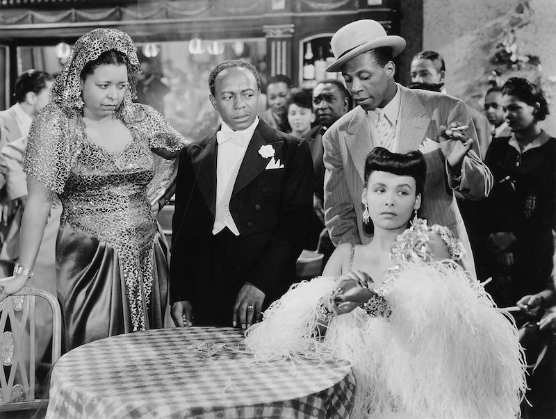 A black and white still from the movie Cabin in the Sky. The stars congreate around a table. A man and woman look incredulously at woman who is seated at the table wearing an evening gown and feather wrap.